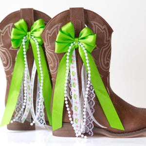 May include: A pair of brown cowboy boots decorated with bright green bows, white lace, and strings of pearls. The boots have decorative stitching and are adorned with ribbons featuring a flamingo pattern.