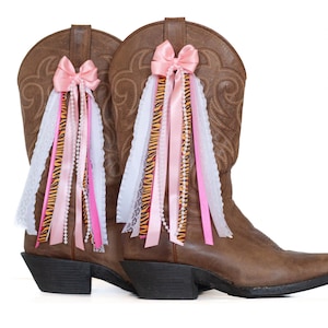 May include: A pair of brown leather cowboy boots adorned with pink bows and cascading ribbons. The ribbons include white lace, pink satin, and beaded strands, with a zebra print accent. The boots have black heels.