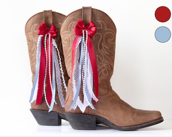 Puede incluir: Un par de botas de vaquero de cuero marrón adornadas con lazos rojos y cintas rojas, blancas y azul claro. Las botas tienen costuras decorativas y un diseño occidental clásico.