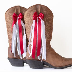 Puede incluir: Un par de botas de vaquero de cuero marrón adornadas con lazos rojos, encaje blanco y hebras de perlas. Las botas tienen un diseño occidental clásico con costuras decorativas y una punta puntiaguda. Los accesorios añaden un toque festivo.
