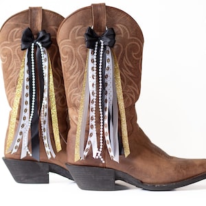 May include: A pair of brown leather cowboy boots adorned with black bows and decorative ribbons. The ribbons include gold glitter, white beaded strands, and ribbons with football designs, creating a festive look.