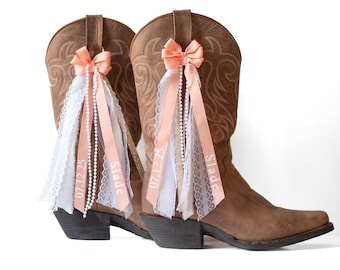 May include: Two brown leather cowboy boots decorated with peach-colored bows, ribbons, and white lace. The ribbons are printed with the name "Slade" and the date "07.12.25". Pearl strands hang from the bows.