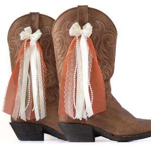May include: A pair of brown leather cowboy boots with decorative accents. Each boot features a white bow and cascading ribbons in white, orange, and cream, adorned with pearls and lace. The boots have a classic western design.