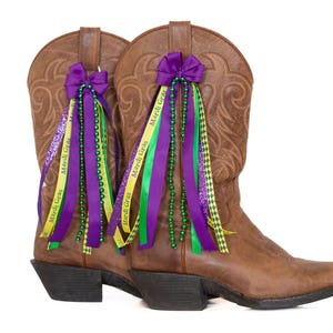 May include: A pair of brown leather cowboy boots decorated with purple bows, ribbons, and green and yellow beads. The ribbons are printed with "Mardi Gras". The boots have a classic western design with pointed toes and stacked heels.