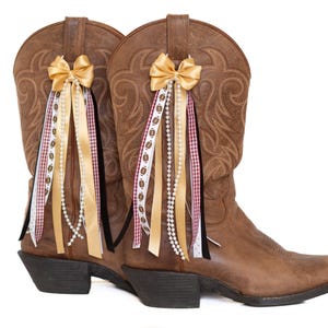 Può includere: Un paio di stivali da cowboy in pelle marrone con fiocchi e nastri decorativi. I fiocchi sono dorati e i nastri includono bianco, nero e bordeaux. Alcuni nastri hanno un design a pallone da football. Gli stivali hanno un classico design western.