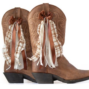 May include: A pair of brown leather cowboy boots with decorative bows, ribbons, and pearl strands. The boots have a classic western design with intricate stitching and a pointed toe. The embellishments include brown bows, white and brown plaid ribbons, and strings of pearls.
