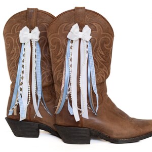 May include: A pair of brown leather cowboy boots with decorative white bows and flowing ribbons in white, light blue gingham, and pearl accents. The boots have a classic western design.