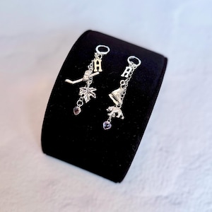 May include: A pair of silver-toned dangle earrings featuring hoop tops. One earring has a letter H charm, a hockey stick, a maple leaf, and a small heart. The other has a letter H charm, an ice skate, a bear figure, and a small heart charm.