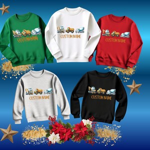 Puede incluir: Cinco sudaderas de cuello redondo en verde, blanco, rojo, gris y negro. Cada sudadera presenta un gr&aacute;fico de veh&iacute;culos de construcci&oacute;n y el texto "CUSTOM NAME" en marr&oacute;n. Las sudaderas se muestran sobre un fondo azul con estrellas doradas y decoraciones florales.