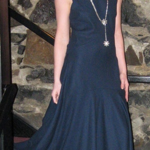 May include: A woman wearing a dark blue dress with a silver chain necklace featuring two flower-shaped pendants. She is standing in front of a stone wall.