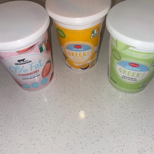Könnte beinhalten: Drei Becher griechischen Joghurts mit weißen Deckeln. Die Geschmacksrichtungen sind Erdbeere, Mango & Passionsfrucht und natürlicher irischer Joghurt. Der Erdbeerbecher ist rosa, der Mango-Becher ist gelb und der Naturjoghurt-Becher ist grün. Jeder Becher ist mit 0 % Fett gekennzeichnet.