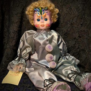 Puede incluir: Una muñeca vintage con la cara pintada y cabello rubio rizado. La muñeca viste un traje de payaso de satén plateado con pompones morados y detalles en negro. La cara tiene diseños florales y coloridos. Una etiqueta dice "Blossomhill".