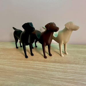 May include: Three Labrador Retriever dog figurines, one black, one brown, and one white, standing on a light wood surface.