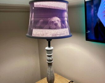 May include: A gray table lamp with a fabric lampshade featuring a photo of a sleeping dog. The lamp has a distressed wood base.