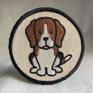 May include: A round embroidered patch featuring a cartoon beagle dog with brown and white fur. The dog is sitting with its tail curled behind it. The patch is surrounded by a black border.