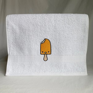 May include: White bath towel with an embroidered yellow popsicle with a bite taken out of it and a smiling face.