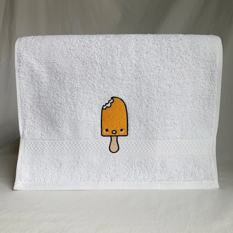 May include: White bath towel with an embroidered yellow popsicle with a bite taken out of it and a smiling face.