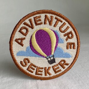 May include: A brown embroidered patch with the words "Adventure Seeker" in a circle around a purple and yellow hot air balloon with blue clouds.
