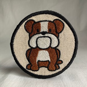 May include: A brown and white embroidered bulldog patch on a beige background with a black circle border.
