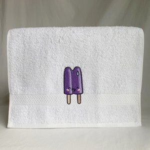 May include: White bath towel with a purple popsicle embroidered on it. The popsicle has a smiling face and a single drop of sweat above its eye.