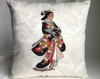 May include: A white pillow with an embroidered image of a woman in a black and red kimono with floral patterns. The woman is standing with her hands clasped in front of her.