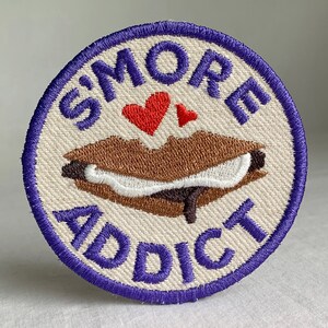 May include: A round, white fabric patch with purple stitching. The patch features the text "S'MORE ADDICT" in purple stitching, a brown and white embroidered s'more, and a red heart.