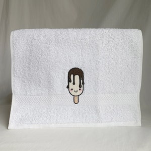May include: White hand towel with a brown and white embroidered ice cream cone with a smiling face.