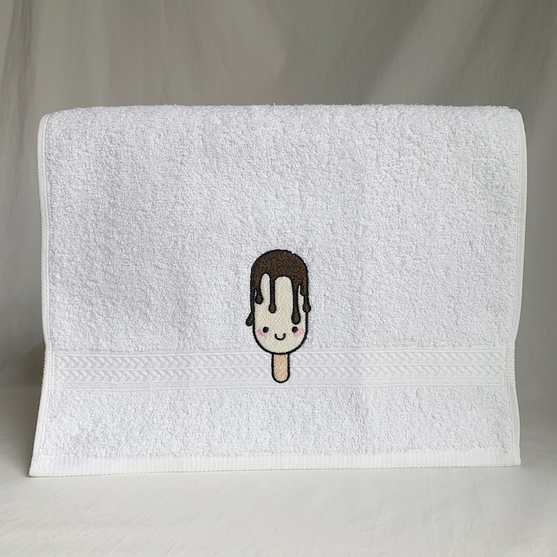 May include: White hand towel with a brown and white embroidered ice cream cone with a smiling face.