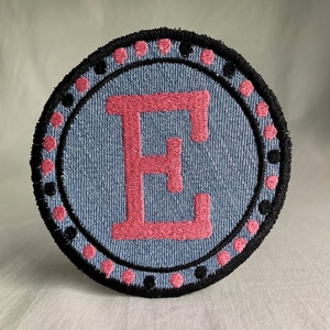 May include: A blue and pink embroidered patch with the letter "E" in the center. The letter is surrounded by a black circle with pink dots.