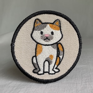 May include: A round, embroidered patch featuring a calico cat with orange, black, and white markings. The cat is sitting with its paws tucked underneath it and has a neutral expression.