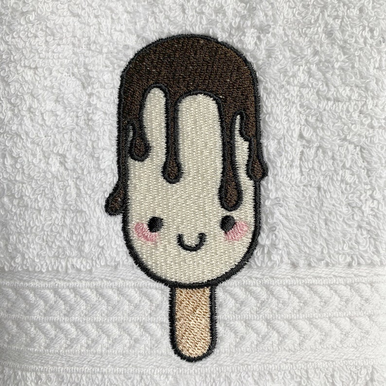 May include: Embroidered white popsicle with a brown chocolate drizzle and a smiling face. The popsicle has a brown stick.