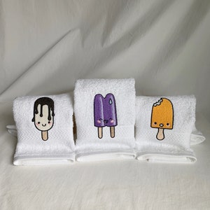 May include: Three white hand towels with embroidered images of popsicles. The popsicles are purple, orange, and chocolate with cute faces.