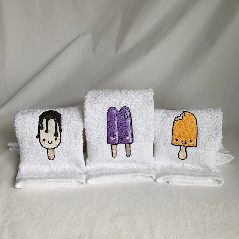 May include: Three white hand towels with embroidered images of popsicles. The popsicles are purple, orange, and chocolate with cute faces.