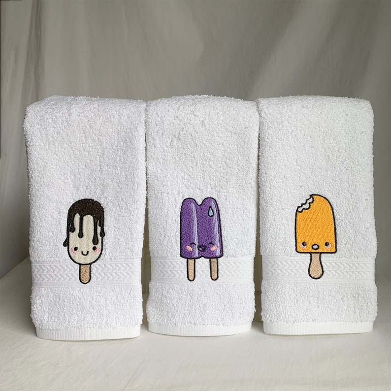 May include: Three white bath towels with embroidered images of popsicles. The popsicles are purple, orange, and chocolate with smiling faces.
