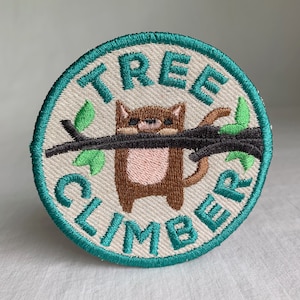 May include: A round, teal embroidered patch with the words "Tree Climber" in white. A brown cartoon animal with pink ears and a tail is hanging from a black branch with green leaves.