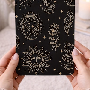 Peut inclure: Une carte rectangulaire noire avec des illustrations dorées de symboles célestes, dont un soleil, un serpent, une fleur et une plume. La carte est tenue par deux mains. L'arrière-plan est flou.