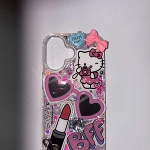 Peut inclure: Coque de téléphone transparente décorée d'un motif Hello Kitty tenant un appareil photo, de lunettes de soleil roses en forme de cœur, d'un rouge à lèvres et des lettres "BFF". L'étui est orné de paillettes roses, de strass et d'un nœud rose.