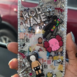 May include: A clear phone case with a glittery background and various embellishments, including a "Rod Wave" logo, a pink and white skull, a black and green robot, and a black and white cartoon figure. The case is decorated with rhinestones and pearls.