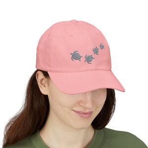 May include: A light pink baseball cap featuring an embroidered design of four sea turtles in a row. The turtles are a light blue color. The cap has a curved brim and a classic six-panel design.