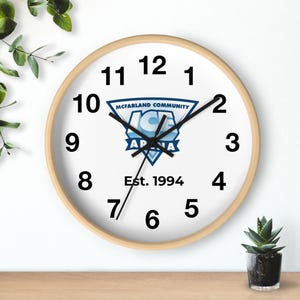 May include: A circular wall clock with a light wood surround. The white face displays black numerals, black hands, and a blue and white logo reading "MCFARLAND COMMUNITY ICE ARENA" and "Est. 1994".