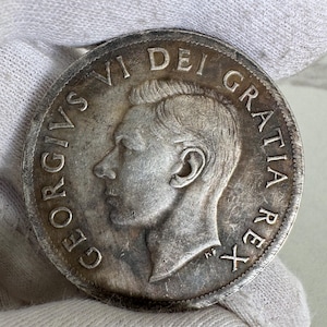 Puede incluir: Una moneda de plata con el perfil de un hombre y la inscripción "GEORGIVS VI DEI GRATIA REX" alrededor del borde. La moneda muestra signos de desgaste y envejecimiento. Se encuentra sobre un fondo blanco.