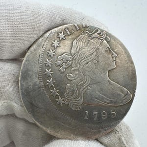 Może przedstawiać: Duża, srebrna moneta z profilem kobiety i rokiem 1795. Moneta ma napis "LIBERTY" i gwiazdy. Moneta jest trzymana przez rękę w rękawicy.