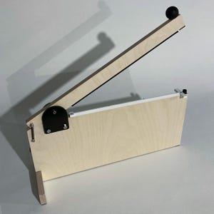 May include: A wooden paper cutter with a light wood finish and black accents. The blade arm is raised, revealing a white cutting edge. A black ball sits at the end of the blade arm. The cutter is designed for precision cutting.