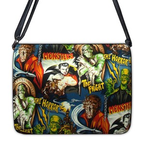 May include: A black shoulder bag with a colorful print of classic monster movie posters. The print features images of Dracula, Frankenstein, the Mummy, and the Wolfman. The bag has a black strap and a zipper closure.
