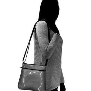 K&ouml;nnte beinhalten: Eine schwarze Umh&auml;ngetasche mit einem silbernen Rei&szlig;verschluss und einem gl&auml;nzenden, strukturierten Finish. Die Tasche wird &uuml;ber der Schulter einer Person getragen.