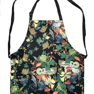 May include: Black apron featuring an owl and floral design. The apron has a black neck strap and ties. The pattern includes owls, leaves, and flowers in green, yellow, orange, and blue. The apron is suitable for cooking or crafting.