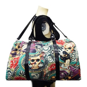 Könnte beinhalten: Bunte Reisetasche mit japanischem Tattoo-Design. Die Tasche zeigt einen lebendigen Druck von Totenköpfen, Schlangen, Blumen und einer Geisha auf einem türkis-violetten Hintergrund. Sie hat schwarze Ledergriffe und einen Schultergurt.