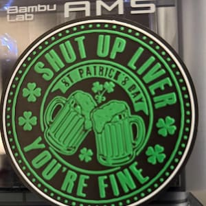 Puede incluir: Un posavasos redondo, negro y verde, con temática del Día de San Patricio. El posavasos presenta el texto "SHUT UP LIVER YOU'RE FINE" alrededor del borde, con dos jarras de cerveza verdes y tréboles en el centro.