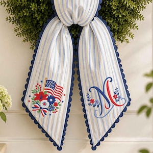 May include: A green wreath with a striped bow in blue and white, trimmed with navy blue scallops. The bow features embroidered details, including an American flag and floral designs. The letter "N" is also embroidered on the bow, adding a personalised touch.