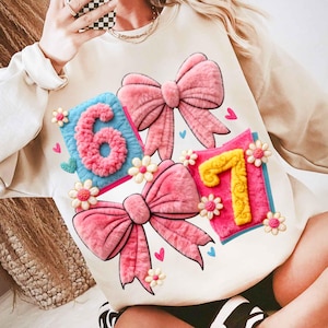 May include: Cream-colored sweatshirt featuring a design with pink bows and the numbers "6" and "7". The numbers are in a fluffy, textured style, set on blue and pink squares. Small flowers and hearts accent the design.